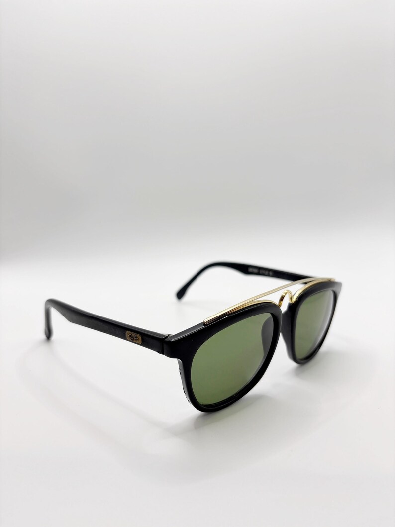 May include: Black sunglasses with green lenses and a gold-colored metal bridge. The sunglasses have a classic design with a rectangular frame and a subtle logo on the temple. The lenses are a dark green color.