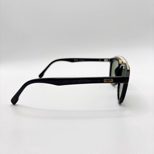 May include: Black sunglasses with a gold accent on the top frame. The lenses are green. The arms are black and curve down to the ear. The sunglasses are set against a white background.