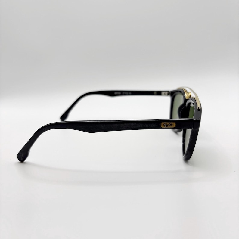 May include: Black sunglasses with a gold accent on the top frame. The lenses are green. The arms are black and curve down to the ear. The sunglasses are set against a white background.