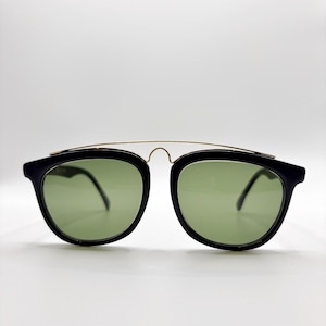 May include: Black sunglasses with green lenses. The sunglasses have a rounded square shape with a gold-colored metal bridge. The frames are black and the lenses are a light green color.