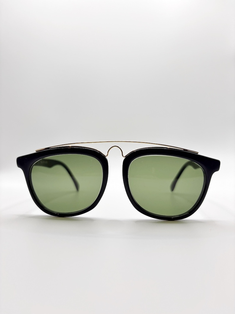 May include: Black sunglasses with green lenses. The sunglasses have a rounded square shape with a gold-colored metal bridge. The frames are black and the lenses are a light green color.