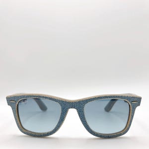 Puede incluir: Gafas de sol azules estilo denim con lentes azul claro. Las monturas tienen un borde de color claro y el logotipo de Ray-Ban es visible en la lente izquierda. Las gafas de sol están sobre un fondo blanco.