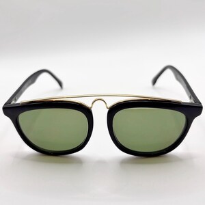 May include: Black sunglasses with a gold-colored bridge and green lenses. The sunglasses have a classic, retro design with a rectangular shape. The frames are black and the lenses are a light green color.