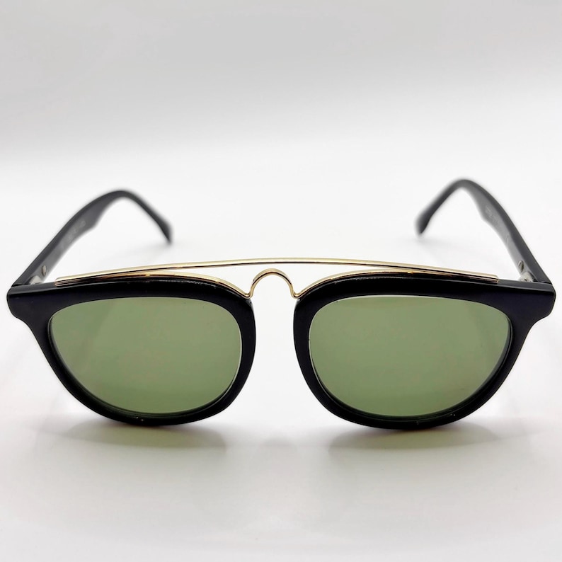May include: Black sunglasses with a gold-colored bridge and green lenses. The sunglasses have a classic, retro design with a rectangular shape. The frames are black and the lenses are a light green color.