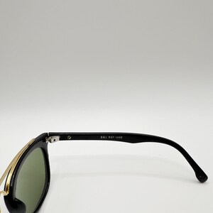 May include: Black sunglasses with green lenses and gold-colored metal accents. The temples are black and feature the text "B&L RAY-BAN". The sunglasses are set against a white background.