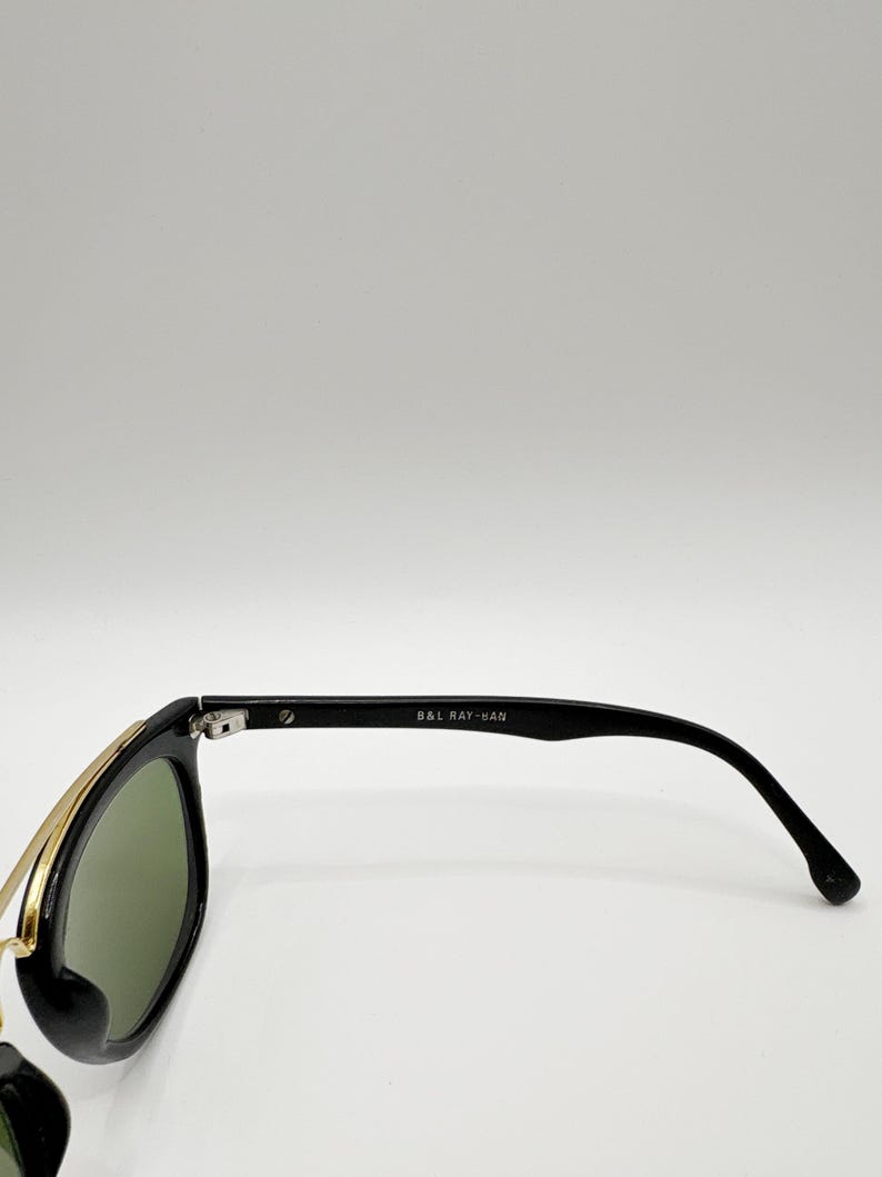 May include: Black sunglasses with green lenses and gold-colored metal accents. The temples are black and feature the text "B&L RAY-BAN". The sunglasses are set against a white background.
