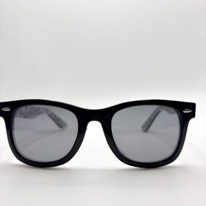 May include: Black sunglasses with grey lenses. The frames are thick and rectangular, with a glossy finish. The temples have a subtle pattern. The sunglasses are set against a white background.