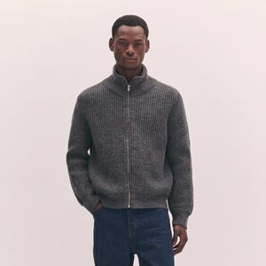 Può includere: Un maglione a coste grigio antracite con collo alto e una cerniera lampo in metallo argentato che corre lungo tutta la parte anteriore. Il capo presenta maniche lunghe con polsini e orlo a coste, realizzato in misto lana.