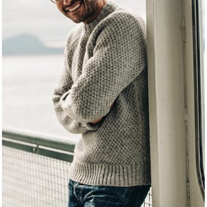 May include: A light grey crew neck jumper featuring a textured waffle knit pattern, long sleeves, and a ribbed hem. The garment is constructed with a thick, woven material and is paired with dark blue denim jeans, showcasing a classic design.