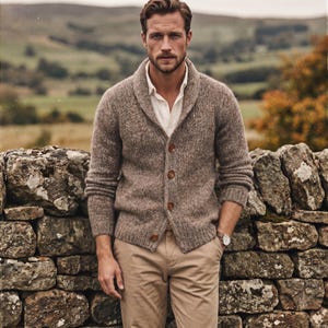 Könnte beinhalten: Eine beige und braun gesprenkelte Strickjacke mit Schalkragen und vier braunen Knöpfen, getragen über einem weißen Hemd mit Kragen und einer beigen Hose. Eine Armbanduhr mit weißem Zifferblatt und braunem Lederarmband ist am linken Handgelenk sichtbar.