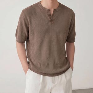 May include: A short-sleeved brown henley shirt featuring a textured knit fabric, a round neckline, and a placket with three matching buttons. The garment has a ribbed hem and ribbed sleeve cuffs, providing a structured fit and a consistent brown tone.