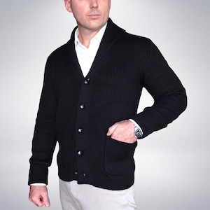 May include: A black ribbed knit cardigan featuring a shawl collar, long sleeves, and a button-front closure. The garment includes two front patch pockets and is worn over a crisp white collared shirt, paired with light-coloured trousers.