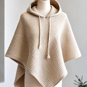 Pode incluir: Um poncho de malha bege com capuz, apresentando um padrão canelado texturizado em todo o tecido. A peça inclui um capuz com dois cordões pendentes a condizer com pequenas extremidades atadas, criando uma silhueta drapeada e assimétrica.