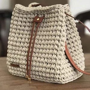 May include: A beige crochet bucket bag featuring a thick, textured knit pattern. The bag includes a brown leather drawstring closure with gold-toned metal accents and a matching brown leather shoulder strap attached to the sides of the bag.