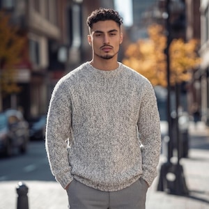 May include: A long-sleeved crew neck sweater featuring a chunky knit texture in a heathered blend of white and grey yarn. The garment has a ribbed collar, cuffs, and hem, providing a structured fit. It is paired with solid grey trousers.