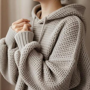 May include: A beige knit hoodie featuring a thick, textured waffle-stitch pattern, a hood with matching drawstrings, and long sleeves with wide, ribbed cuffs. The garment has a relaxed fit with dropped shoulders and a substantial, chunky knit texture.