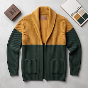 Könnte beinhalten: Eine farblich abgesetzte Strickjacke mit Schalkragen, bestehend aus einem senfgelben Oberteil und einem dunkelwaldgrünen Unterteil. Das Strickkleidungsstück verfügt über zwei aufgesetzte Vordertaschen und ein braunes Lederetikett mit der Aufschrift Moss & Fell.