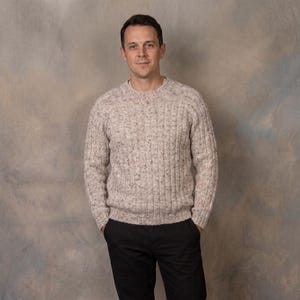 May include: A beige crew neck sweater featuring a vertical ribbed knit pattern with small dark brown and black flecks throughout the fabric, paired with solid black trousers. The sweater has long sleeves and a standard fit with a ribbed hem and cuffs.