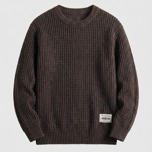 May include: A dark brown long-sleeve sweater featuring a waffle knit texture, a crew neckline, and ribbed cuffs and hem. A small rectangular white fabric patch with red and black text is stitched onto the lower left side of the garment.