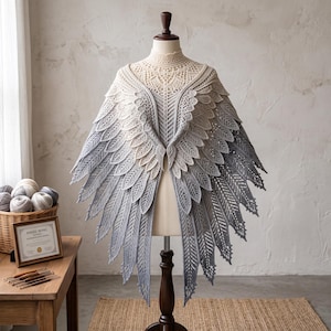 May include: A crochet shawl featuring an intricate wing-like design with a gradient color transition from cream at the neckline to dark gray at the pointed edges. The garment displays detailed feather-like patterns and a high-neck collar structure.