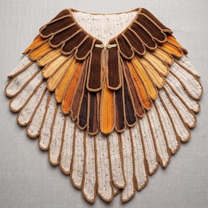 May include: A crochet capelet featuring a layered, feather-like design in shades of dark brown, orange, and cream with speckled details. The garment is secured at the neckline with a gold-toned metal clasp and consists of multiple rows of elongated, rounded panels that create a textured, wing-inspired appearance.
