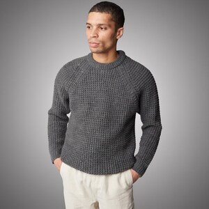 May include: A charcoal grey long-sleeve jumper featuring a thick waffle-knit texture, a round crew neckline, and raglan sleeves. The jumper is paired with off-white trousers, creating a neutral-toned ensemble with a visible knit pattern.
