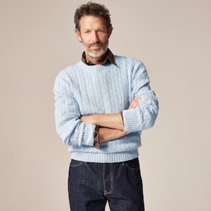 Puede incluir: Un suéter de punto trenzado en color azul claro con cuello redondo y mangas largas con puños acanalados, usado sobre una camisa de cuello marrón oscuro, combinado con pantalones vaqueros de color azul oscuro.