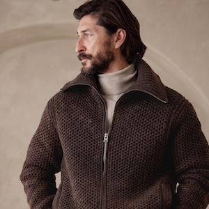 May include: A dark brown, chunky knit zip-up sweater featuring a wide, ribbed collar and a silver-toned metal zipper closure. The sweater is worn over a light beige, high-neck turtleneck base layer, creating a layered, textured appearance.
