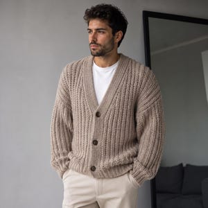 May include: A beige chunky knit cardigan featuring a deep V-neckline, long sleeves with ribbed cuffs, and three dark brown buttons down the front. The garment is worn over a plain white t-shirt and paired with light-colored trousers.