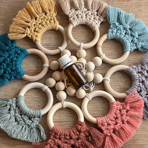 May include: A selection of colourful macrame baby teethers in shades of yellow, beige, teal, grey, and coral. Each teether has a wooden ring and a decorative fringe. A small brown essential oil bottle and wooden beads are in the centre.