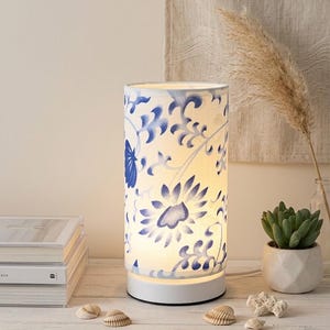 Peut inclure: Lampe de table cylindrique avec une base blanche et un abat-jour orné d'un motif floral bleu. La lampe est allumée, dégageant une douce lueur. Des coquillages décoratifs et une petite plante succulente en pot sont à proximité.
