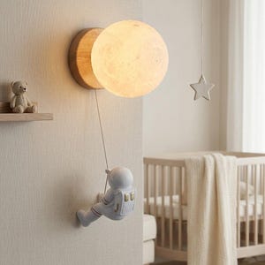 Peut inclure: Lampe murale décorative en forme de lune éclairée, fixée sur un support en bois. Une petite figurine d'astronaute semble descendre de la lune. Parfait pour une chambre d'enfant, cette lampe est un élément décoratif.