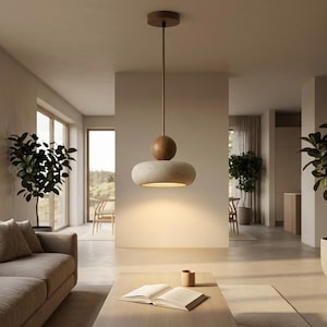 Puede incluir: Una moderna lámpara colgante con una parte superior de madera y una pantalla redondeada beige. La luz cuelga de un cable marrón e ilumina una sala de estar con una paleta de colores neutros. La habitación incluye un sofá, plantas y una mesa de centro.