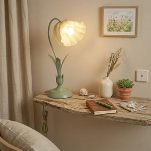Peut inclure: Une lampe de table avec une base en métal vert sauge en forme de tige de fleur et un abat-jour en verre blanc dépoli rappelant une fleur épanouie. La lampe est allumée, diffusant une lueur chaude, avec un col incurvé et des feuilles.