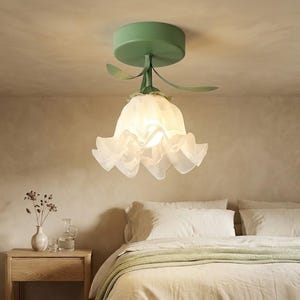 May include: A ceiling-mounted light fixture featuring a sage green circular base and a stem with two leaf-shaped accents. The shade is crafted from translucent, white, ruffled glass that mimics the appearance of a blooming flower petal shape.