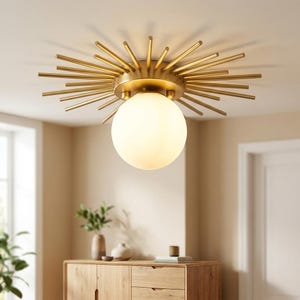 May include: A ceiling light fixture featuring a central frosted white glass globe surrounded by a sunburst-style frame made of thin, radiating metallic gold rods that extend outward from a circular base, creating a starburst effect.