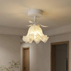 May include: A white, flower-shaped ceiling light. The light fitting has a white base and a translucent, petal-like shade. The light is mounted on a white ceiling, illuminating the room. The room has wooden doors.