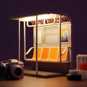 May include: A miniature model of a New York City subway car interior. The model features orange seating, a poster of the Statue of Liberty, and a lit overhead panel. The overall colour scheme is beige, orange, and white.