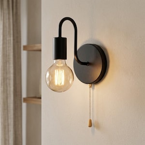 Könnte beinhalten: Schwarze Wandleuchte mit einer großen, runden, klaren Glühbirne. Die Leuchte hat einen gebogenen Arm und eine Zugkette mit einem Holzschalter. Das Licht ist auf einer kreisförmigen schwarzen Basis montiert.