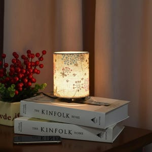 Può includere: Una piccola lampada cilindrica con paralume a fantasia floreale, appoggiata su due libri intitolati "The Kinfolk Home". La lampada emette un bagliore caldo, illuminando il motivo floreale nelle tonalità del rosa, blu e beige. Sullo sfondo una piccola pianta con bacche rosse.