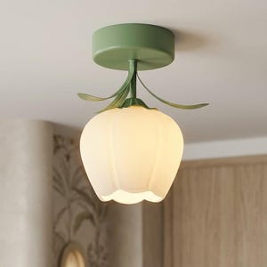 Könnte beinhalten: Eine Deckenleuchte mit grünem Sockel und floralen Details. Die Leuchte hat einen weißen, tulpenförmigen Schirm. Sie ist an einer weißen Decke montiert, mit einer gemusterten Wand und einer Holztür im Hintergrund. Ein dekoratives Wohnaccessoire.