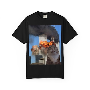 Op de afbeelding: Zwart t-shirt met een meme van een kat met grote ogen voor de brandende Twin Towers. De afbeelding is geplaatst tegen een blauwe lucht met rook en vlammen. De kat is bruin en wit.