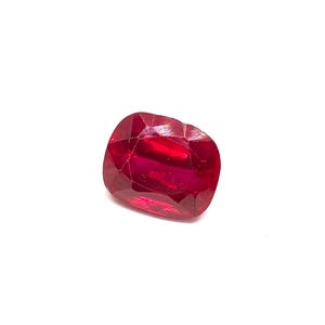 May include: A faceted, cushion-cut ruby gemstone. The gem is a deep, vibrant red colour, with visible internal inclusions. The shape is a rounded rectangle, against a white background.