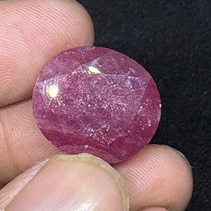 May include: A faceted, oval-shaped ruby gemstone. The ruby is a deep pinkish-red color, with visible internal inclusions and a polished surface. The gemstone is held between fingers against a dark background.