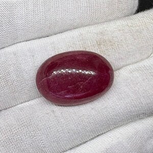 Può includere: Una gemma lucida, di forma ovale, di colore rosso intenso. La gemma ha una superficie liscia e riflettente, su uno sfondo di tessuto bianco. Il colore della pietra è una tonalità ricca e satura, che suggerisce possa essere un rubino.