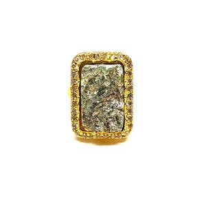 May include: A gold-toned ring with a rectangular, textured, green and gray stone center. The stone is surrounded by a border of small, clear stones. The ring has a simple band.