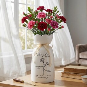 May include: A white ceramic vase with a scalloped top, filled with red and pink carnations. The vase has a cream-colored ribbon and text that reads "Chapter Fifty". A wooden table holds the vase, a letter opener, and books.