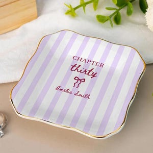 May include: A decorative ceramic ring dish with a gold rim and a striped lavender and white pattern. The dish has the text "CHAPTER thirty" and "Amelia Smith" in burgundy. A silver ring with a diamond is next to the dish.