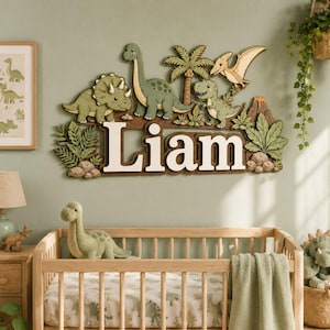 May include: A wall-mounted wooden sign featuring the name Liam in large white letters, surrounded by layered cutouts of green dinosaurs, a palm tree, a flying pterodactyl, and a volcano, all set against a muted sage green wall background.