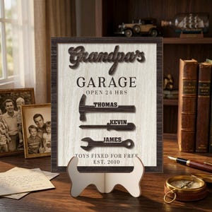 May include: Wooden sign with the words "Grandpa's Garage" and the names Thomas, Kevin, and James. The sign includes the text "Toys Fixed For Free" and "Est. 2010". The sign is displayed on a wooden stand.